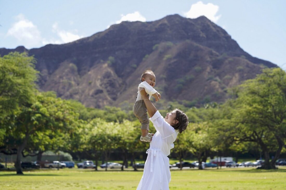 青空の下、ハワイのダイヤモンドヘッドを背景にした緑豊かなカピオラニ公園で、母親が満面の笑みを浮かべながら子どもを高く抱き上げている様子。